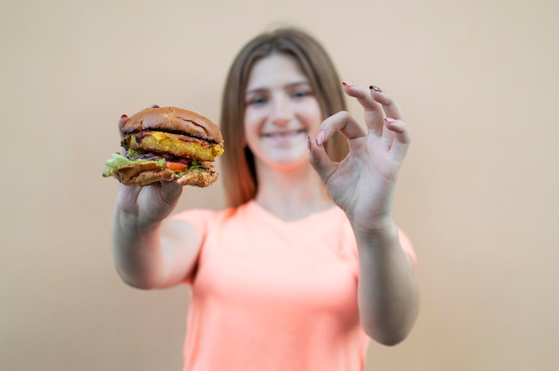 Aantrekkelijk tienermeisje houdt een grote hamburger vast en laat met haar hand zien dat het goed is