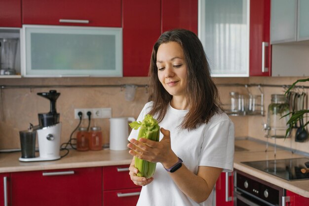 Aantrekkelijk vrolijk meisje staat in de keuken met verse groenten en selderij in haar handen te houden