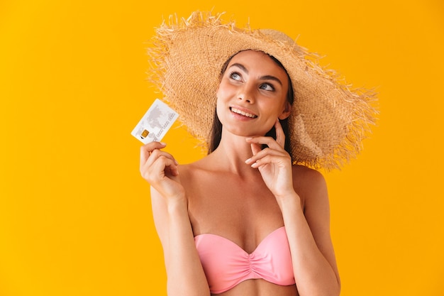 Aantrekkelijk vrolijk peinzend jong meisje met een bikini die over een gele muur staat en een plastic creditcard toont