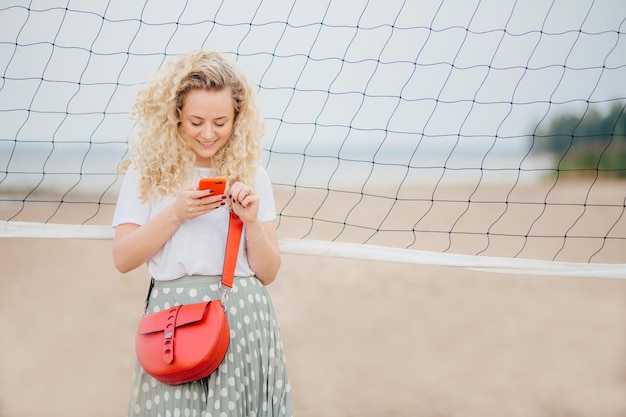 Aantrekkelijk vrouwelijk model gericht op smartphone leest informatie op webpagina heeft een heldere tas glimlacht vreugdevol als bericht ontvangen van vriendje poses op strand Mensen levensstijl en rust concept