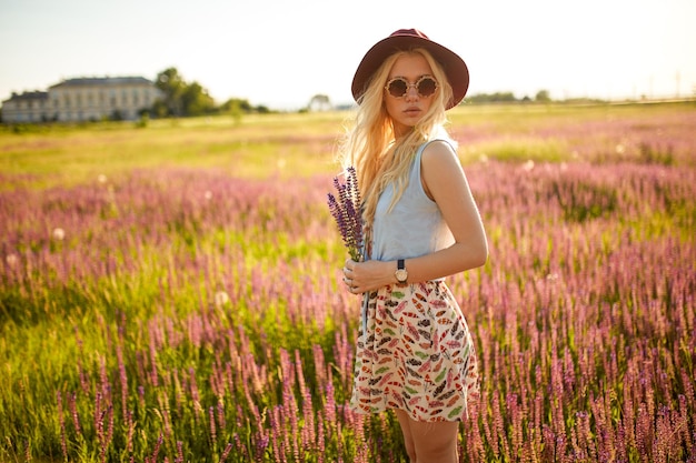 Aantrekkelijk vrouwelijk model met blond haar draagt een hoed en een ronde zonnebril die poseert in een bloemenveld en een zonsondergangachtergrond