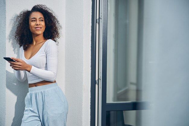 Aantrekkelijke Afro-Amerikaanse vrouw die in lichte kleding tegen de muur staat en haar mobiel vasthoudt