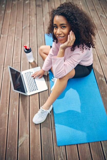 Aantrekkelijke Afro-Amerikaanse vrouw die op yogamat zit en droomt terwijl ze online op laptop werkt