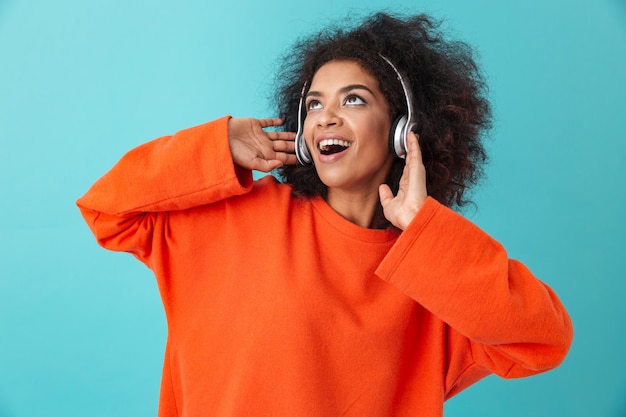 Aantrekkelijke Amerikaanse vrouw in oranje shirt genieten van muziek via draadloze oortelefoons terwijl u luistert naar favoriete melodie, geïsoleerd over blauwe muur