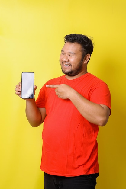 Aantrekkelijke Aziatische dikke man in rode tshirt wijst vinger naar leeg scherm smartphone geïsoleerd op yel
