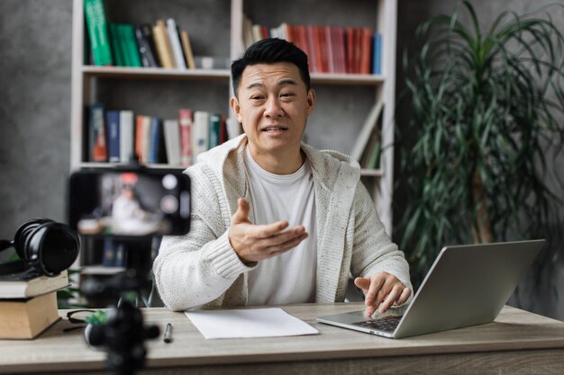 Aantrekkelijke aziatische man die op laptop werkt en video opneemt op digitale smartphone