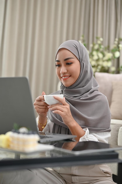 Aantrekkelijke Aziatische moslimvrouw die koffie drinkt tijdens het gebruik van een laptop