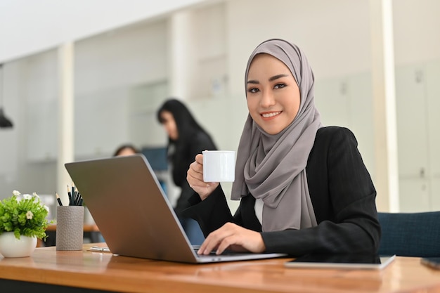 Aantrekkelijke Aziatische moslimzakenvrouw die hijab draagt en 's ochtends koffie drinkt op kantoor