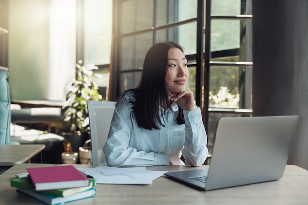 Aantrekkelijke aziatische onderneemster die aan laptop op modern kantoor werkt en wegkijkt