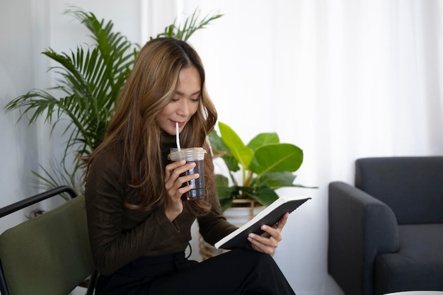 Aantrekkelijke aziatische vrouw die een boek leest en ijskoffie drinkt in de woonkamer