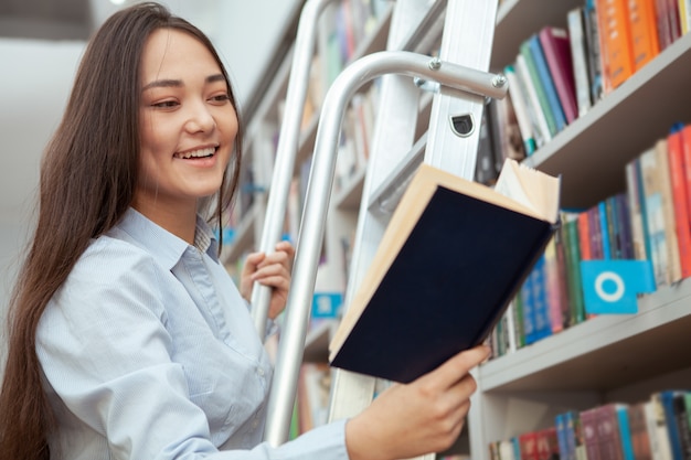 Aantrekkelijke Aziatische vrouw die lacht, een boek leest in de bibliotheek