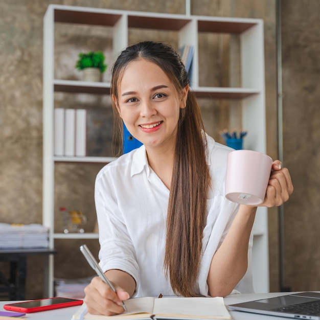 Aantrekkelijke Aziatische vrouw die werkt met een laptop met een koffiekopje op kantoor aan huis