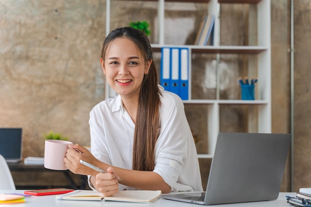 Aantrekkelijke Aziatische vrouw die werkt met een laptop met een koffiekopje op kantoor aan huis