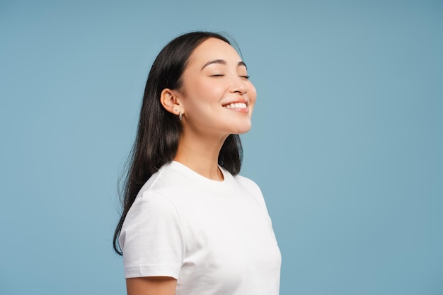 Aantrekkelijke Aziatische vrouw geniet van frisse lucht met gesloten ogen die geïsoleerd staat op een blauwe achtergrond