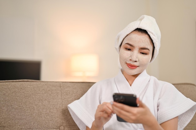 Aantrekkelijke Aziatische vrouw in badjas die haar mobiele telefoon gebruikt om te chatten terwijl ze een gezichtsmasker aanbrengt