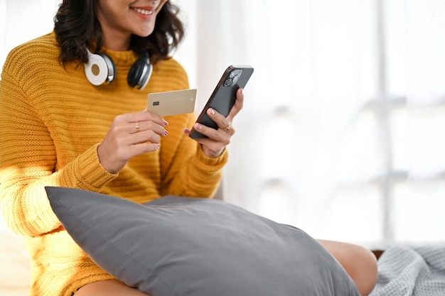 Aantrekkelijke Aziatische vrouw zittend op haar bed met haar smartphone en creditcard bijgesneden