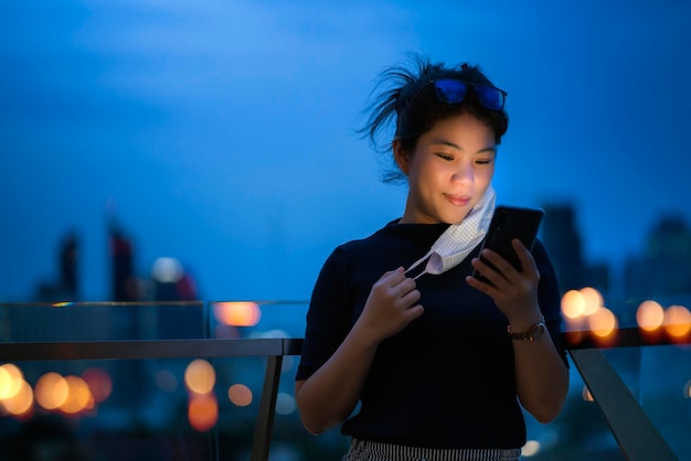 Aantrekkelijke Aziatische vrouwelijke vrouw dragen beschermend masker hand gebruik smartphone technologie communicatie met achtergrond van bokeh licht stedelijk gebouw achtergrond zakelijke ideeën concept