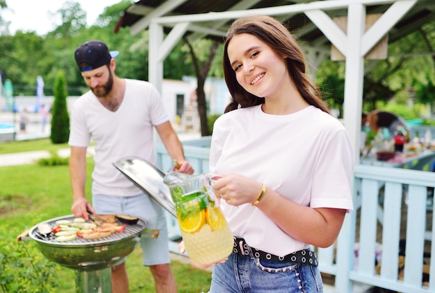 Foto aantrekkelijke bebaarde man frituurt vlees in een barbecue, een jong meisje houdt een glazen kan met verse limonade vast