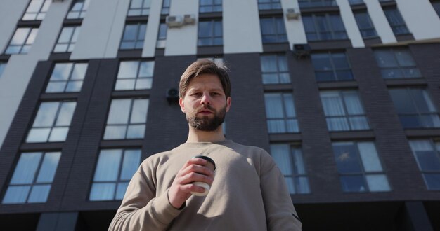 Aantrekkelijke bebaarde man met afhaalbeker met koffie en kijkt recht in de camera op straat