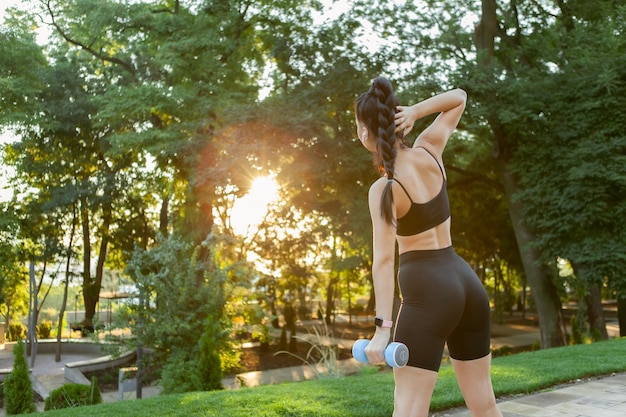 Aantrekkelijke blanke brunette vrouw die laterale buikspieren traint met een halter in haar hand in het park bij zonsopgang