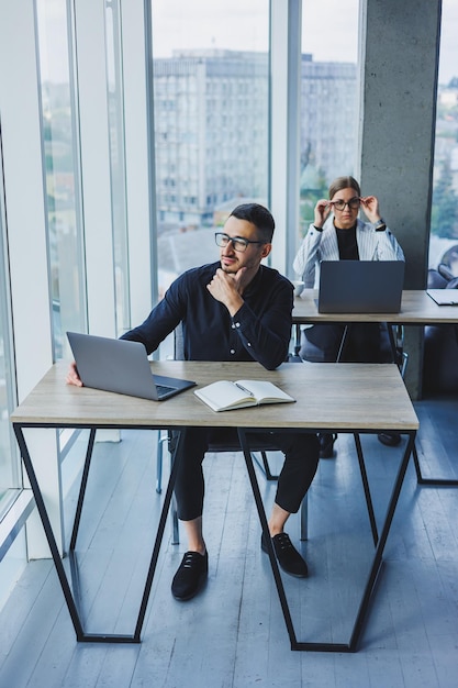 Aantrekkelijke blanke zakenman in een zwart overhemd zit aan een bureau en gebruikt een laptop in een modern kantoor met collega's op de achtergrond Werksfeer op kantoor