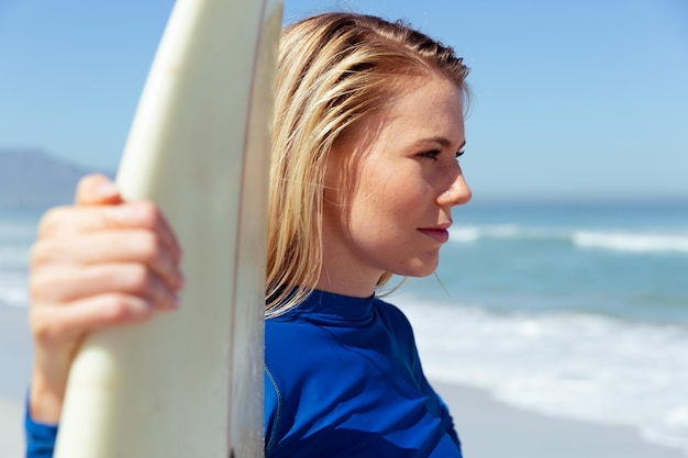 Foto aantrekkelijke blonde blanke vrouw geniet van de tijd op het strand op een zonnige dag, met een surfplank in haar hand en glimlachend, met blauwe hemel en zee op de achtergrond. zomer tropische strandvakantie.