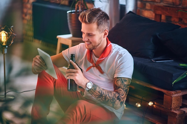 Aantrekkelijke blonde man met tatoeages op armen, gekleed in een rode kerstbroek met tablet-pc in een kamer met loft-interieur.