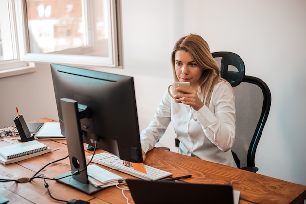 Aantrekkelijke blonde onderneemster die met computer en het drinken koffie in bureau werken.