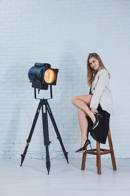 Aantrekkelijke blonde slanke vrouw in een zwarte jurk en schoenen met hoge hakken zit op een stoel in de studio en poseert