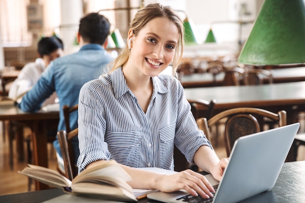 Aantrekkelijke blonde studente studeert in de bibliotheek met laptopcomputer