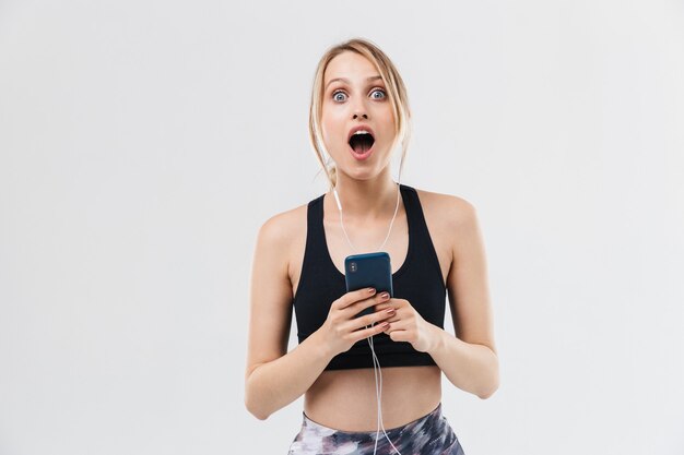 Aantrekkelijke blonde vrouw gekleed in sportkleding trainen en luisteren naar muziek met smartphone tijdens fitness in sportschool geïsoleerd over witte muur