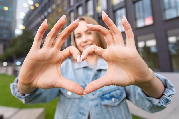 Foto aantrekkelijke blonde vrouw toont haar hart met haar handen draagt een casual jean jas staan in de buurt van een moderne