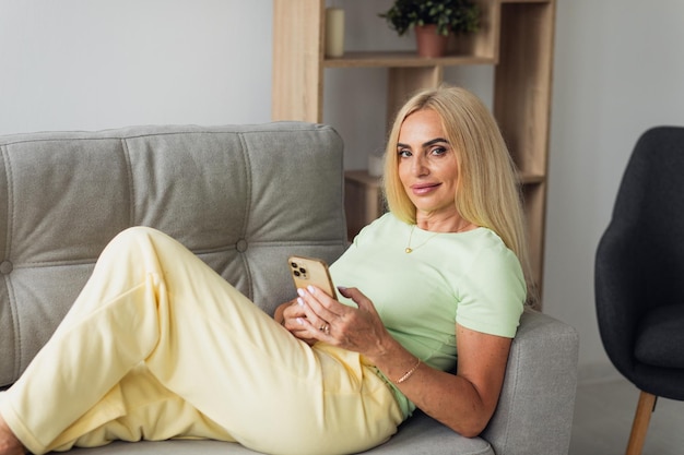 Aantrekkelijke blonde vrouw van middelbare leeftijd gebruikt telefoon terwijl ze thuis op de bank in de woonkamer zit