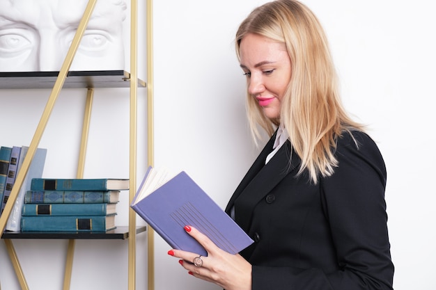 Aantrekkelijke blonde zakenvrouw in een pak staat bij de boekenplank en leest een boek