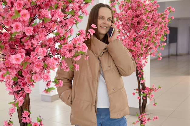 Foto aantrekkelijke bruinharige blanke vrouw met beige bovenkleding die poseert in een winkelcentrum bij prachtige roze bloemen die met een smartphone praat en met een vriend praat.