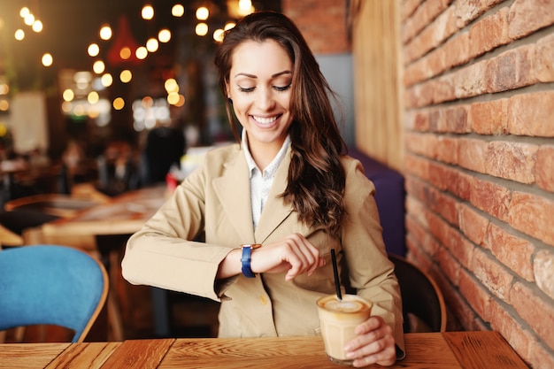 Aantrekkelijke brunette met brede glimlach gekleed smart casual kijken naar horloge en koffie drinken in de cafetaria.