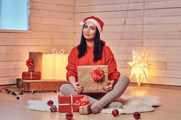 Aantrekkelijke brunette vrouw in een rode kleding zit op een vloer en houdt kerstcadeaus in een kamer met witte houten muur.