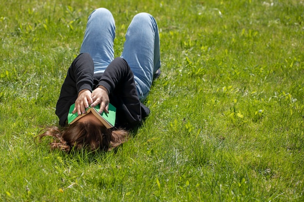 Aantrekkelijke brunette vrouw met lang haar gekleed in zwarte hoodie liggend op gras van groen gazon op mooie zomerdag die betrekking hebben op haar gezicht met boek