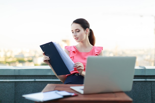 Aantrekkelijke businesslady in roze blouse zit op het dak en werkt met laptop