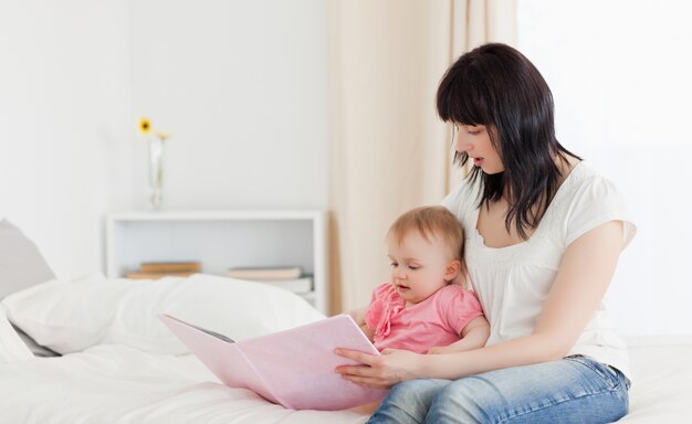 Aantrekkelijke donkerbruine vrouw die een boek toont aan haar baby terwijl het zitten op een bed