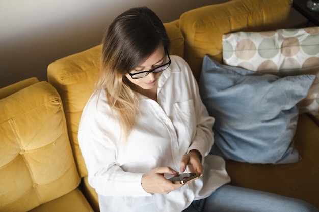 Aantrekkelijke donkerbruine vrouw van middelbare leeftijd met een bril die een mobiele telefoon gebruikt die op een gele bank zit