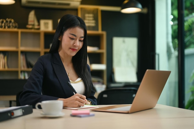 Aantrekkelijke duizendjarige vrouwelijke ondernemer die laptop gebruikt en haar businessplan op notitieblok schrijft