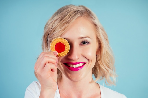 Aantrekkelijke en lachende vrouw blonde met make-up en lippenstift op lippen kleur fuchsia houdt een calorierijk koekjeskoekje in de hand in de studio op een blauwe achtergrond