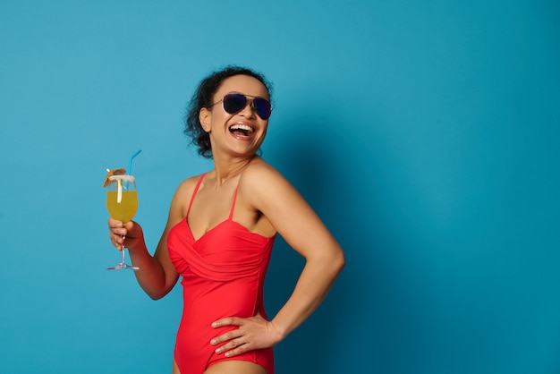 Aantrekkelijke fit vrouw in rode zwembroek en zonnebril met een cocktail op blauwe achtergrond