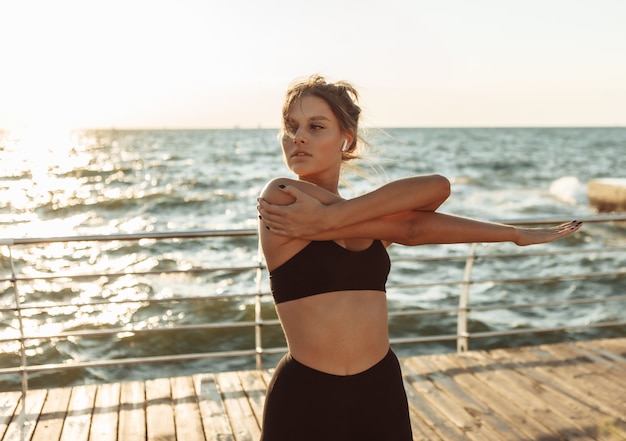 Aantrekkelijke fitte vrouw in sportkleding die de hand uitrekt voordat ze bij zonsopgang op het strand gaat trainen