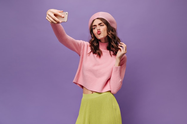 Aantrekkelijke gekrulde vrouw in groene rok, roze baret en roze trui blaast kus, neemt selfie en houdt telefoon op paarse muur