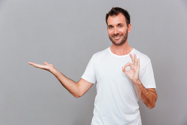 Aantrekkelijke gelukkig lachende casual jonge man in wit t-shirt met copyspace op palm en ok gebaar tonen over grijze achtergrond