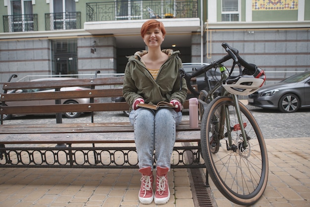 Aantrekkelijke gelukkige jonge vrouw die vreugdevol lacht, zittend op een bankje dichtbij haar fiets