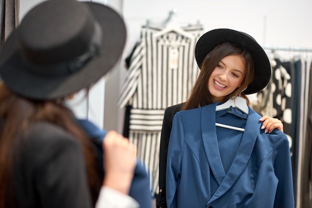 Aantrekkelijke gelukkige jonge vrouw glimlachend vreugdevol terwijl het passen van nieuwe kleren in de modewinkel poseren voor de spiegel copyspace lifestyle mensen positiviteit welzijn consumentisme concept.