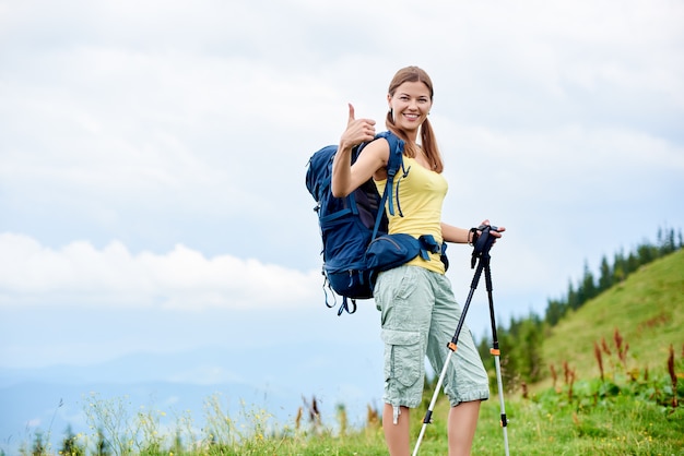 Aantrekkelijke gelukkige vrouw die backpacker de bergsleep wandelen, op grasrijke heuvel lopen, trekkingsstokken gebruiken, aan de camera glimlachen en duimen tonen. Outdoor-activiteit, toerisme concept